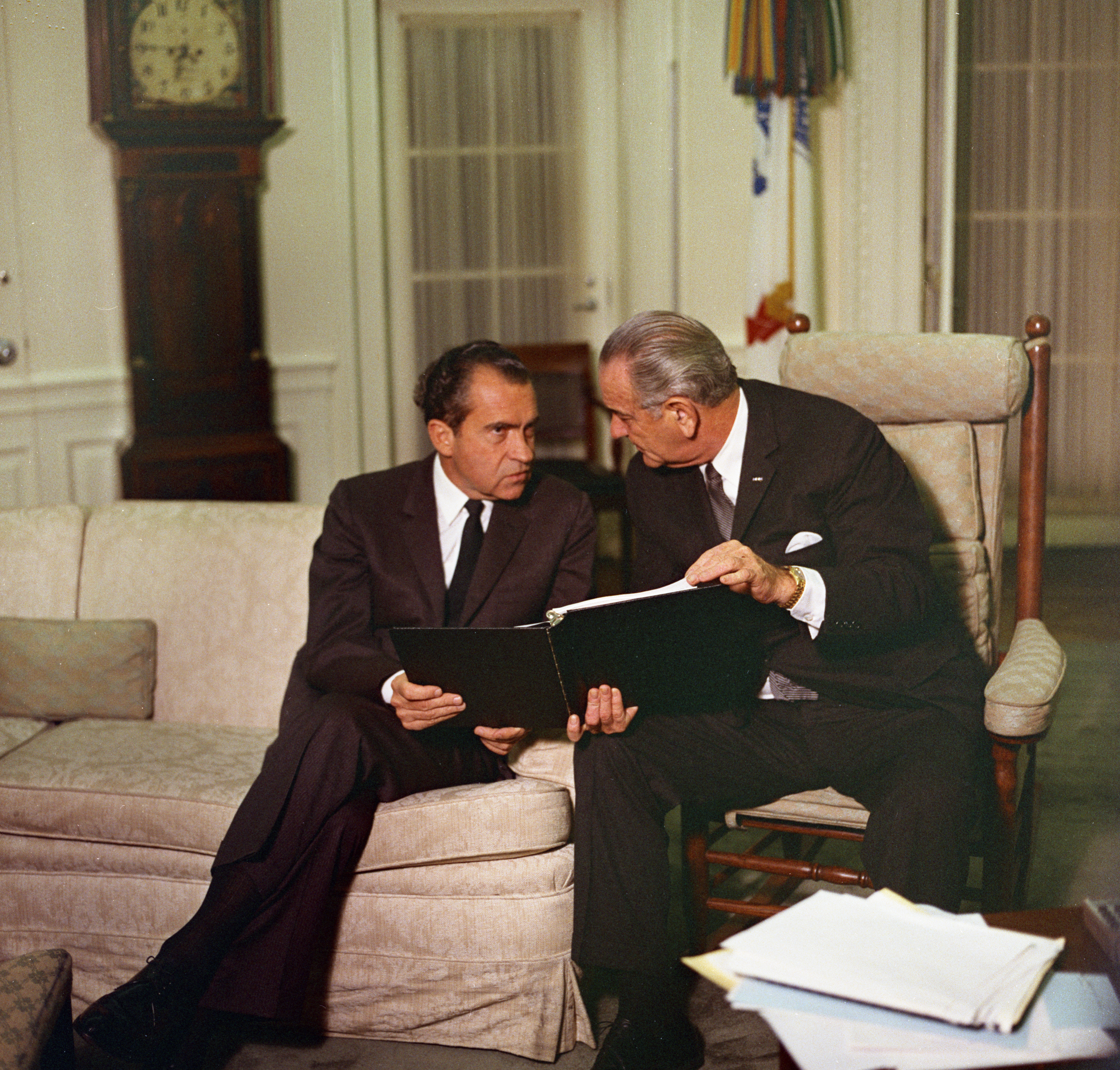 Richard Nixon sit on a sofa and confers with President Lyndon B. Johnson, who sits in a chair next to him and holds a binder. 